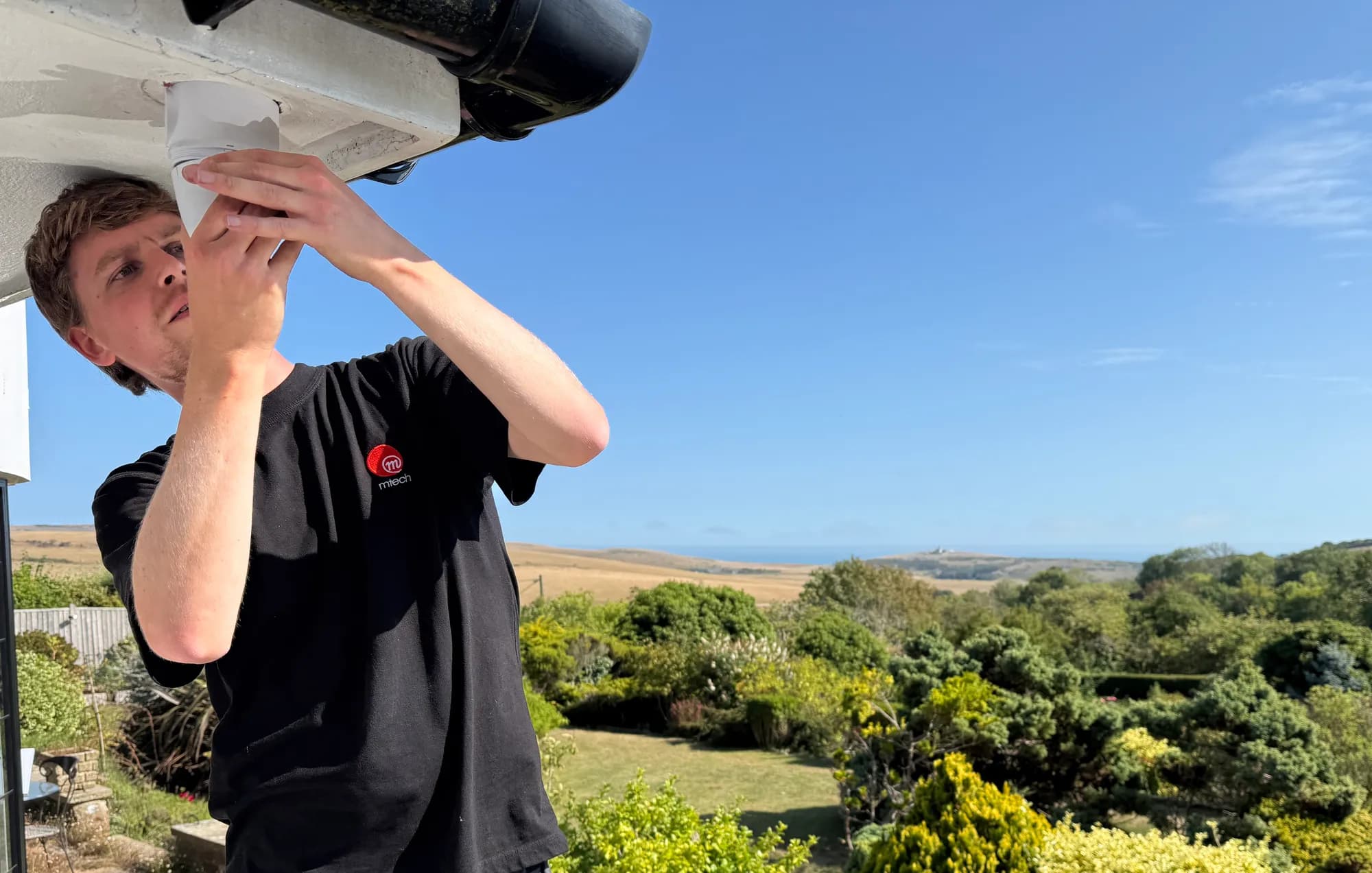 M-Tech engineer fitting a UniFi Protect camera under the eaves of a house overlooking the South Downs