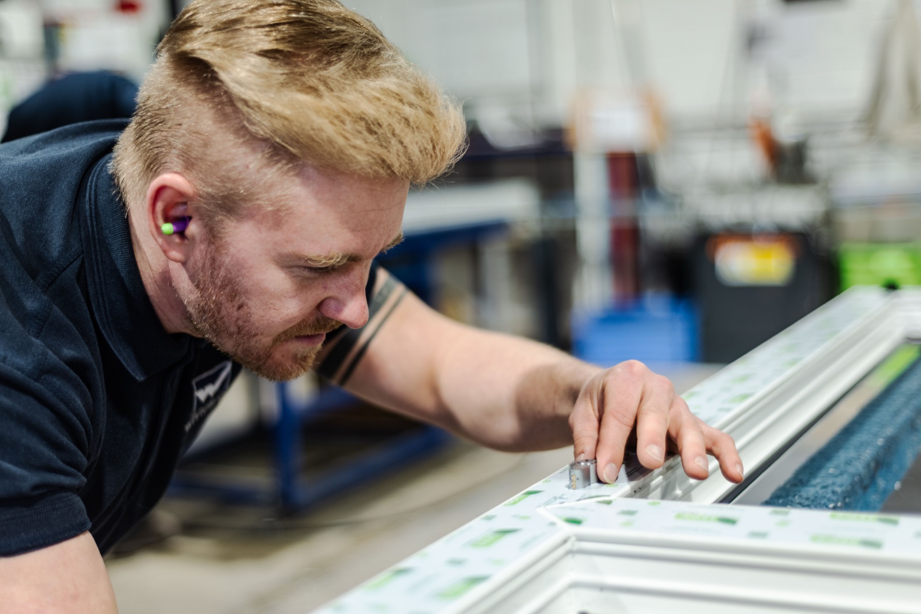 Whiteline production technician in branded polo and ear protection fitting hardware to a uPVC window frame on the workshop bench